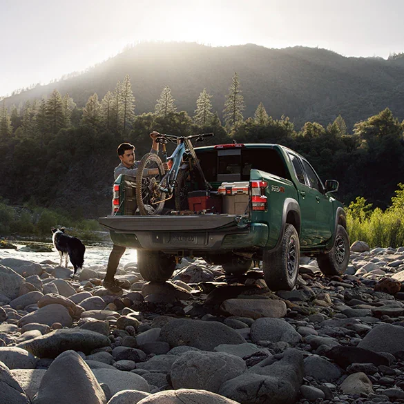 Nissan Frontier parked on a rocky stream shore; the driver is adjusting a bicycle and cargo in the back, while his dog is looking at the stream